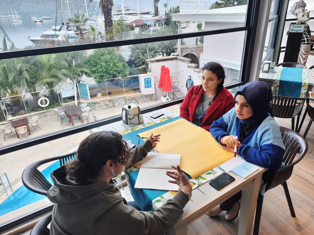 3 katılımcı atölye çalışması için masa etrafında oturuyor ve masada renkli kartonlar var