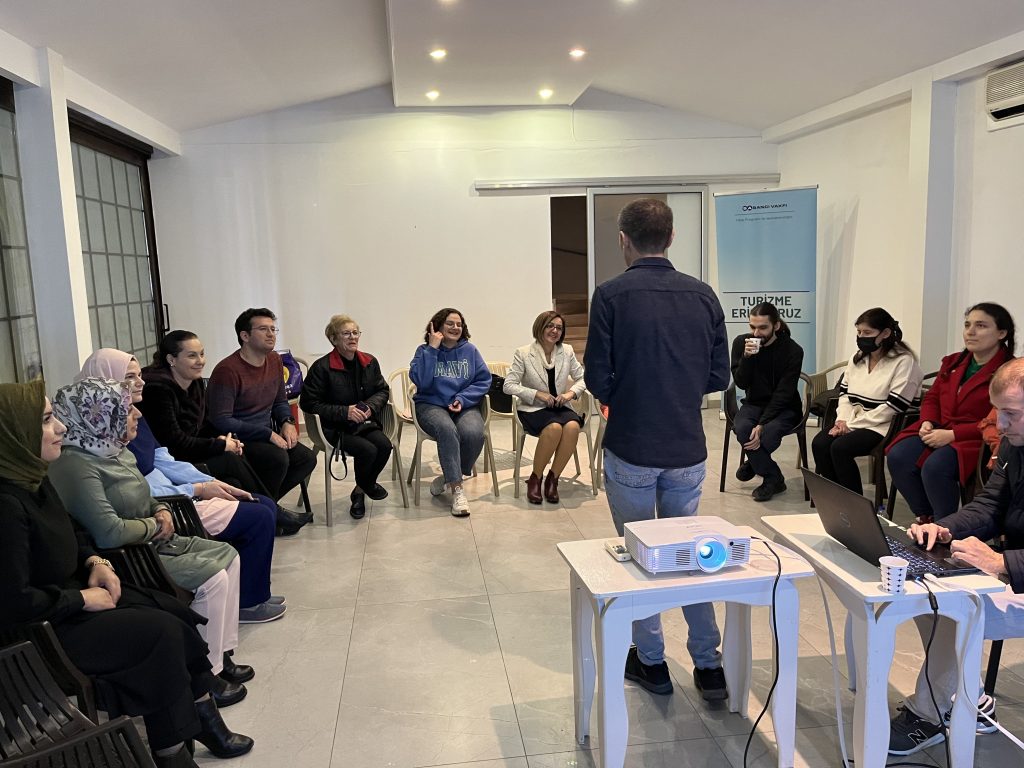 Kameraya sırtını dönerek 8-10 kişiye yüz yüze eğitim vermekte olan erkek eğitmen