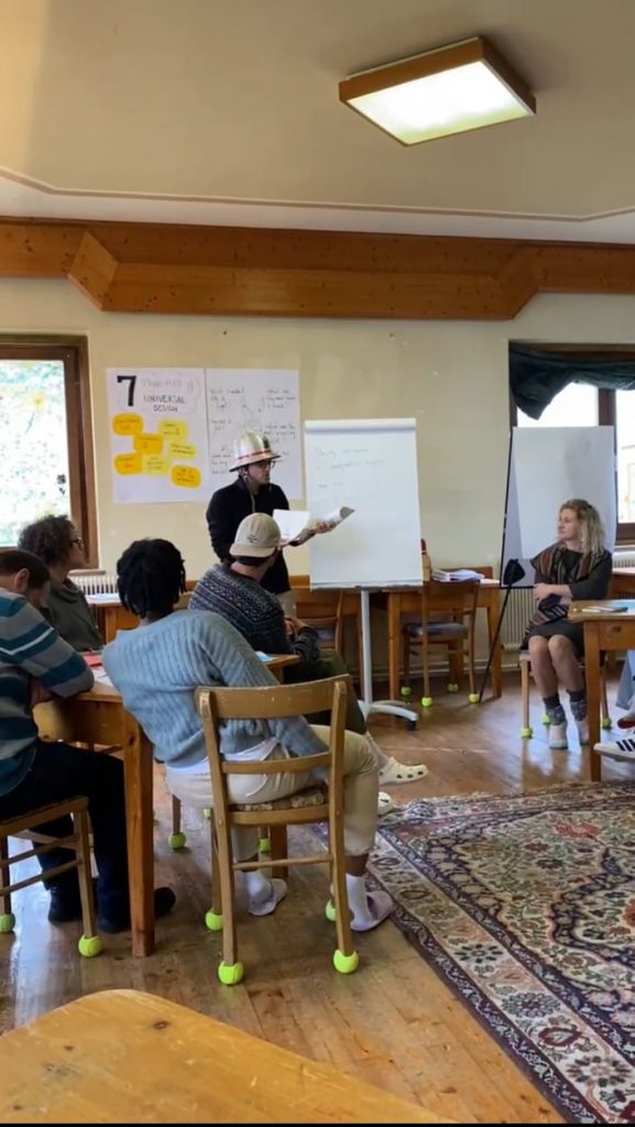 Sahnede sunum yapan bir katılımcıyı izlemekte olan katılımcılar yer almakta.