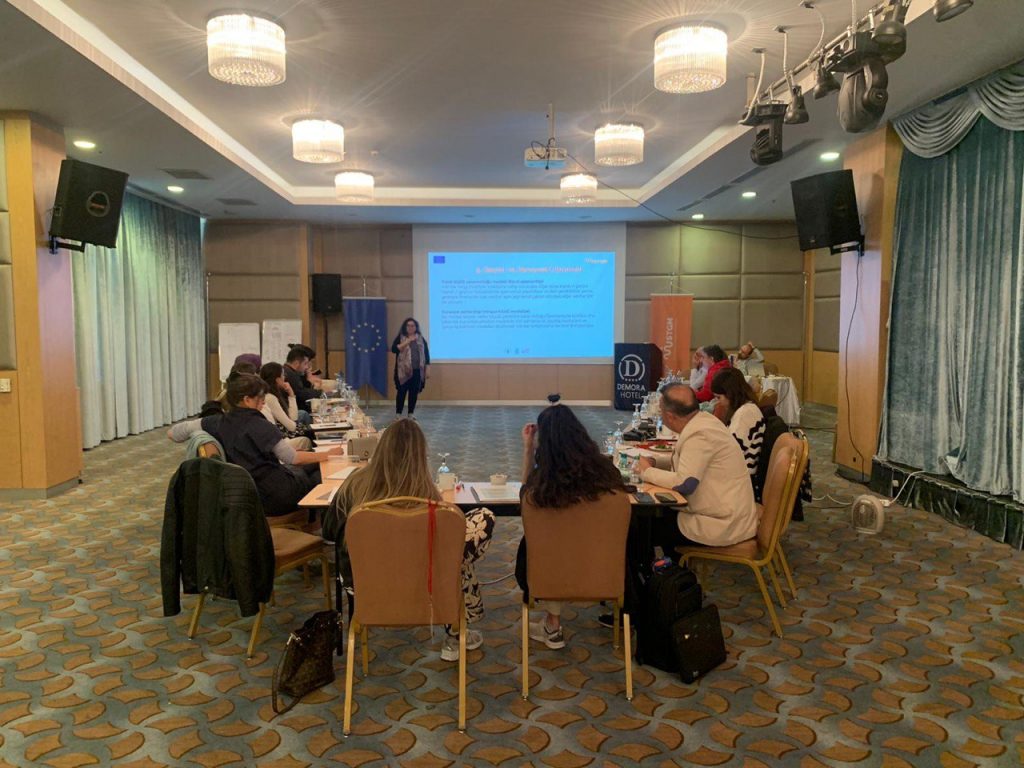 Projeksiyona yansıtılmış eğitim materyalinin önünde U düzeninde masada oturan 20 kişi var