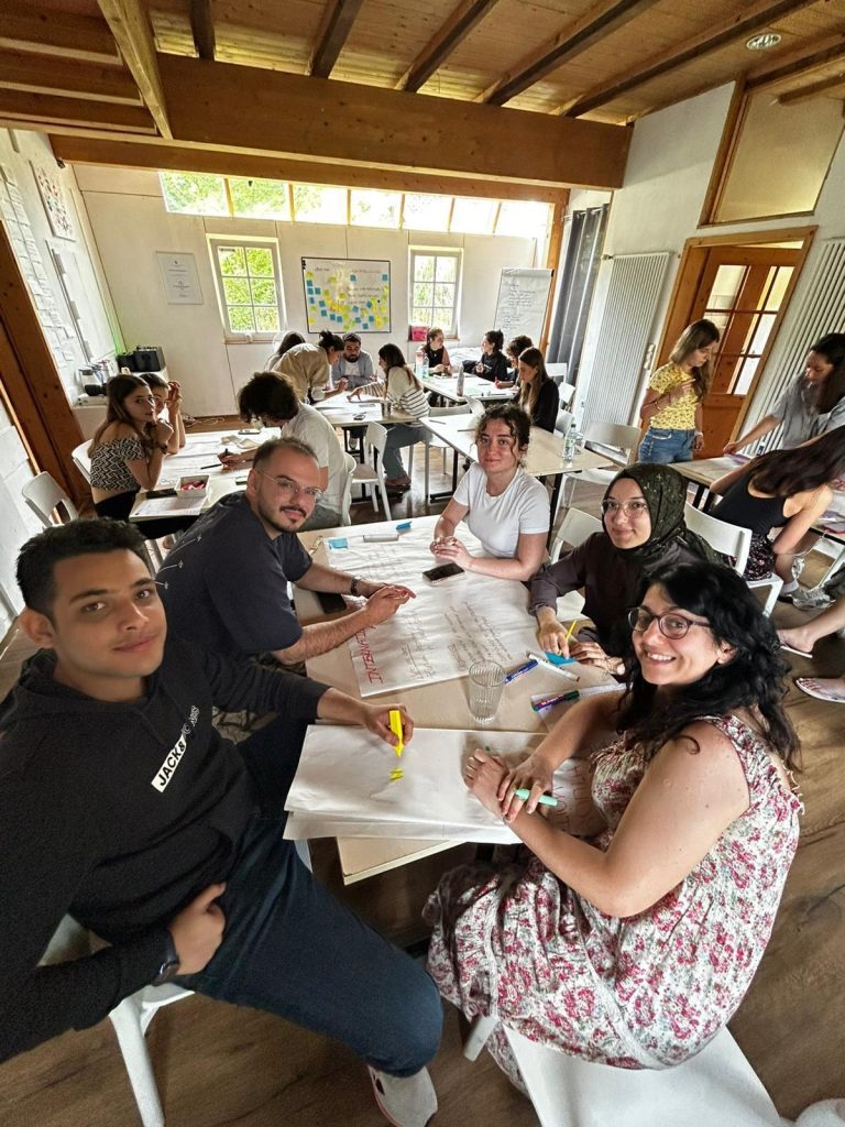 Beş kişiden oluşan bir grup, kapalı bir odada yan yana ayakta duruyor ve birlikte ellerinde büyükçe bir afiş tutuyorlar. Arka planda beyaz tahta ve not kağıtlarıyla dolu bir pano yer alıyor. Odanın tavanı eğimli ve üst kısmı camlarla kaplı; dışarıdan yeşil ağaçlar görünüyor. Işık doğal olarak içeriye süzülüyor.
Soldan sağa:
İlk kişi, siyah tişört ve desenli krem renkli pantolon giymiş. Kısa saçlı ve sakallı. Sağ eliyle afişin sol üst köşesini tutuyor.
İkinci kişi, siyah kapüşonlu bir sweatshirt ve koyu renk pantolon giymiş. Afişi ortasından tutuyor.
Üçüncü kişi, siyah başörtüsü takıyor, kahverengi tunik ve beyaz spor ayakkabılar giymiş. Afişi orta alt bölümünden destekliyor.
Dördüncü kişi, beyaz tişört ve açık mavi kot pantolon giymiş. Afişe dokunmuyor.
Beşinci kişi, çiçek desenli beyaz-pembe bir elbise giymiş ve gözlük takıyor. Kollarını göğsünde birleştirmiş, gülümsüyor.
Afiş Üzerinde:
Büyük harflerle "INTERNATIONAL YOUTH QUEST ASSOCIATION" yazıyor. Alt kısmında Türkiye bayrağı çizilmiş. Ayrıca engelsiz yaşam, engelli bireylerin sosyal hayata katılımı, kapsayıcılık gibi konuları içeren İngilizce ifadeler yer alıyor. Sol alt köşede bir tekerlekli sandalye simgesi bulunuyor.