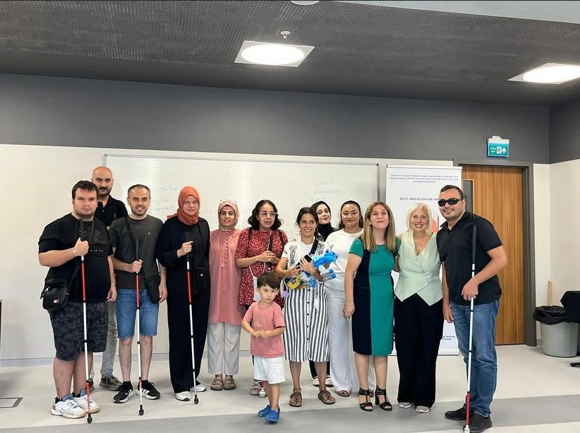 Fotoğrafta bir grup insan kapalı bir ortamda, muhtemelen bir eğitim salonunda veya toplantı odasında bir araya gelmiş. Arka planda beyaz bir yazı tahtası ve bir bilgilendirme afişi bulunuyor. Grupta kadınlar, erkekler ve bir çocuk yer alıyor. Katılımcıların bazıları beyaz baston taşıyor; bu, görme engelli bireylerin de bulunduğunu gösteriyor. Herkes birbirine yakın duruyor ve gülümseyerek poz veriyor. Ortam sıcak, samimi ve birlikte geçirilen anlamlı bir etkinliğin ardından çekilmiş gibi görünüyor.