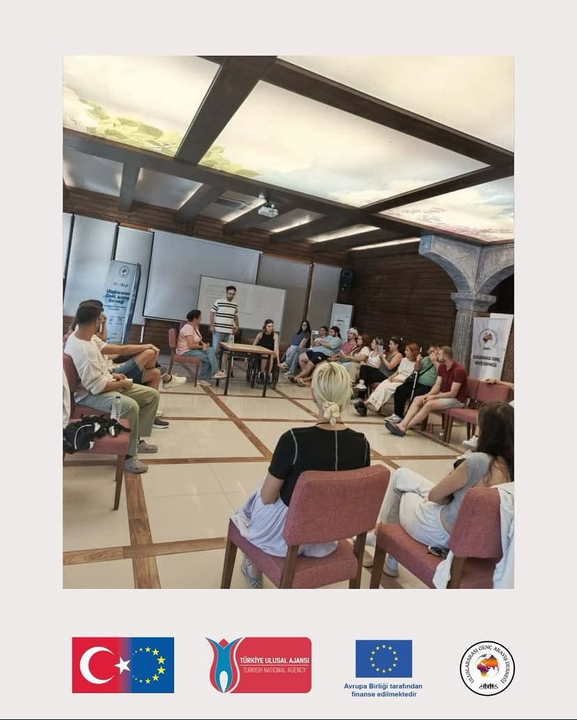 Fotoğrafta bir grup insan kapalı bir toplantı salonunda yarım daire biçiminde oturmuş şekilde görülüyor. Ortada iki kişi ayakta duruyor, biri konuşuyor ya da sunum yapıyor gibi görünüyor. Katılımcılar dikkatle dinliyor. Mekân ahşap detaylara sahip, tavanında doğa temalı bir ışıklandırma paneli bulunuyor. Ortam sıcak, doğal ve samimi bir atmosfer yaratıyor. Arka planda beyaz bir tahta, projeksiyon perdesi ve bir afiş yer almakta. Fotoğrafın alt kısmında dört logo bulunuyor: Türkiye ve Avrupa Birliği bayraklarının birleşimi, Türkiye Ulusal Ajansı logosu, Avrupa Birliği tarafından finanse edilmektedir ibaresi içeren AB logosu, ve Uluslararası Genç Arayış Derneği logosu. Bu detaylardan, görselin Erasmus+ programı kapsamında düzenlenen bir gençlik çalışması ya da eğitim etkinliği sırasında çekildiği anlaşılıyor. Genel atmosfer etkileşimli, katılımcı ve öğrenmeye açık bir grup çalışması havasında.