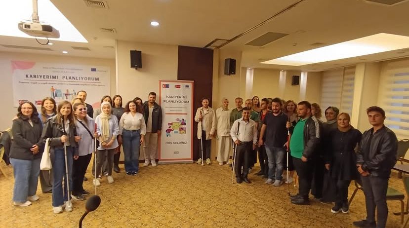 Fotoğrafta geniş bir toplantı salonunda bir grup insan bir araya gelmiş şekilde görülüyor. Grup, fotoğraf için bir araya toplanmış ve kameraya doğru gülümseyerek poz veriyor. Kadınlar ve erkeklerden oluşan katılımcıların bir kısmı beyaz baston taşıyor; bu, görme engelli bireylerin de etkinlikte yer aldığını gösteriyor. Arka planda iki afiş dikkat çekiyor. Afişlerde ve duvardaki yazıda “Kariyerimi Planlıyorum” ifadesi yer alıyor. Afişlerin üst kısmında Türkiye ve Avrupa Birliği bayrakları ile Türkiye Ulusal Ajansı logosu bulunuyor. Bu da etkinliğin Avrupa Birliği destekli bir proje kapsamında gerçekleştirildiğini düşündürüyor.
