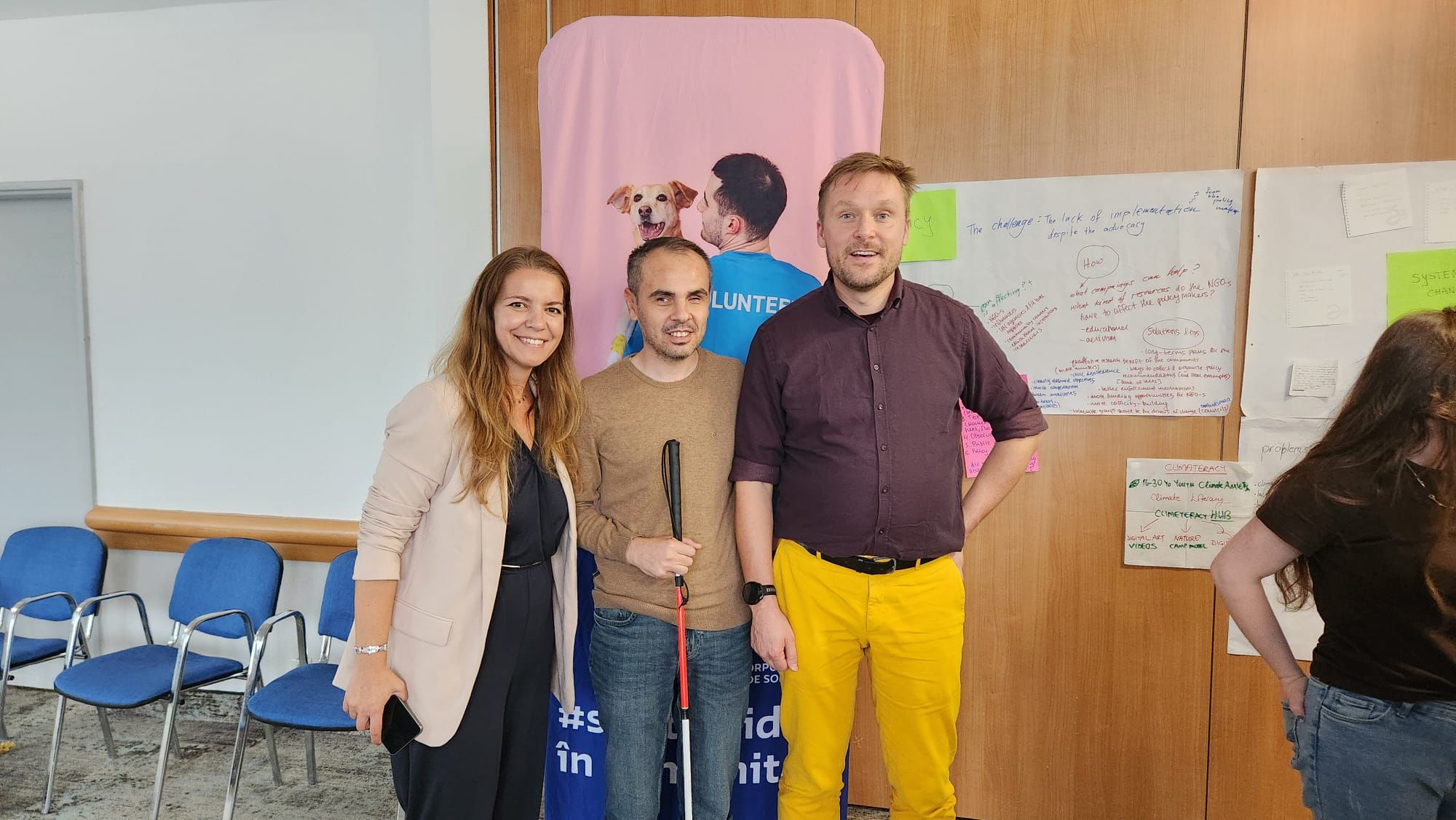 Fotoğrafta üç kişi ön planda yan yana duruyor ve gülümsüyor. Arka planda bir duvarda çeşitli renklerde not kağıtları ve üzerinde yazılar olan büyük kâğıtlar asılmış. Ayrıca ortada pembe bir pano üzerinde köpekli bir görsel bulunuyor. Soldaki kadın uzun saçlı, açık renk bir ceket ve koyu renk bir kıyafet giymiş, elinde telefon tutuyor. Ortadaki erkek kahverengi bir kazak ve kot pantolon giymiş, elinde beyaz baston var. Sağdaki erkek koyu renk bir gömlek ve dikkat çekici sarı pantolon giymiş. Fotoğraf, bir atölye, seminer veya gönüllülük etkinliği ortamında çekilmiş gibi görünüyor. Arkadaki yazılardan “volunteer” (gönüllü) ve proje notları dikkat çekiyor.