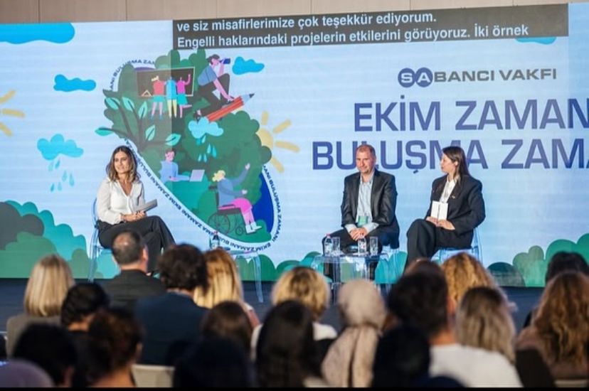 Fotoğrafta bir panel veya toplantı oturumu görülüyor. Arka planda büyük bir ekran üzerinde “Sabancı Vakfı – Ekim Zamanı, Buluşma Zamanı” yazısı yer alıyor. Ekranda ayrıca çeşitli figürlerin yer aldığı, doğa temalı ve kapsayıcılığı vurgulayan bir illüstrasyon bulunuyor — bu illüstrasyonda tekerlekli sandalyedeki bir kişi, bilgisayar başında çalışan gençler ve doğa unsurları dikkat çekiyor. Sahnenin önünde üç kişi oturuyor; biri moderatör konumunda, diğer ikisi konuşmacı olarak görünüyor. İzleyici kitlesi de sahnenin karşısında dikkatle oturmuş durumda. Etkinlik genel olarak profesyonel, kapsayıcı ve ilham verici bir atmosferde gerçekleşiyor. Üst kısımda yer alan altyazıda, konuşmacının engelli haklarına yönelik projelerin etkilerinden söz ettiği görülüyor. Bu fotoğraf, Sabancı Vakfı’nın toplumsal farkındalık ve engelli haklarına dair yürüttüğü projelerin bir yansıması niteliğinde.