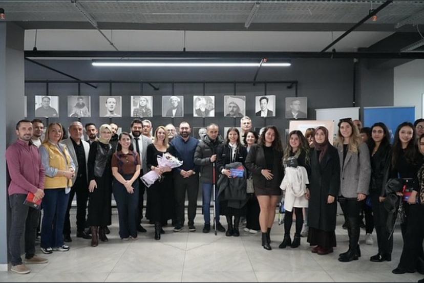 Fotoğrafta, kapalı bir mekânda geniş bir grup insan bir araya gelmiş ve toplu poz vermekte. Grup, yaklaşık 25 kişiden oluşuyor ve ortada çiçek buketi tutan bir kişi dikkat çekiyor. Katılımcılar farklı yaş gruplarından ve çoğunluğu gülümseyerek kameraya bakıyor. Arka planda gri tonlarda bir duvar bulunuyor ve duvarda siyah-beyaz portre fotoğrafları asılı. Üst kısımda modern aydınlatmalar yer almakta. Mekân genel olarak sade, modern ve profesyonel bir ortam izlenimi veriyor. Bu fotoğraf, bir eğitim, toplantı veya proje etkinliğinin sonunda çekilmiş bir hatıra pozu gibi görünüyor.