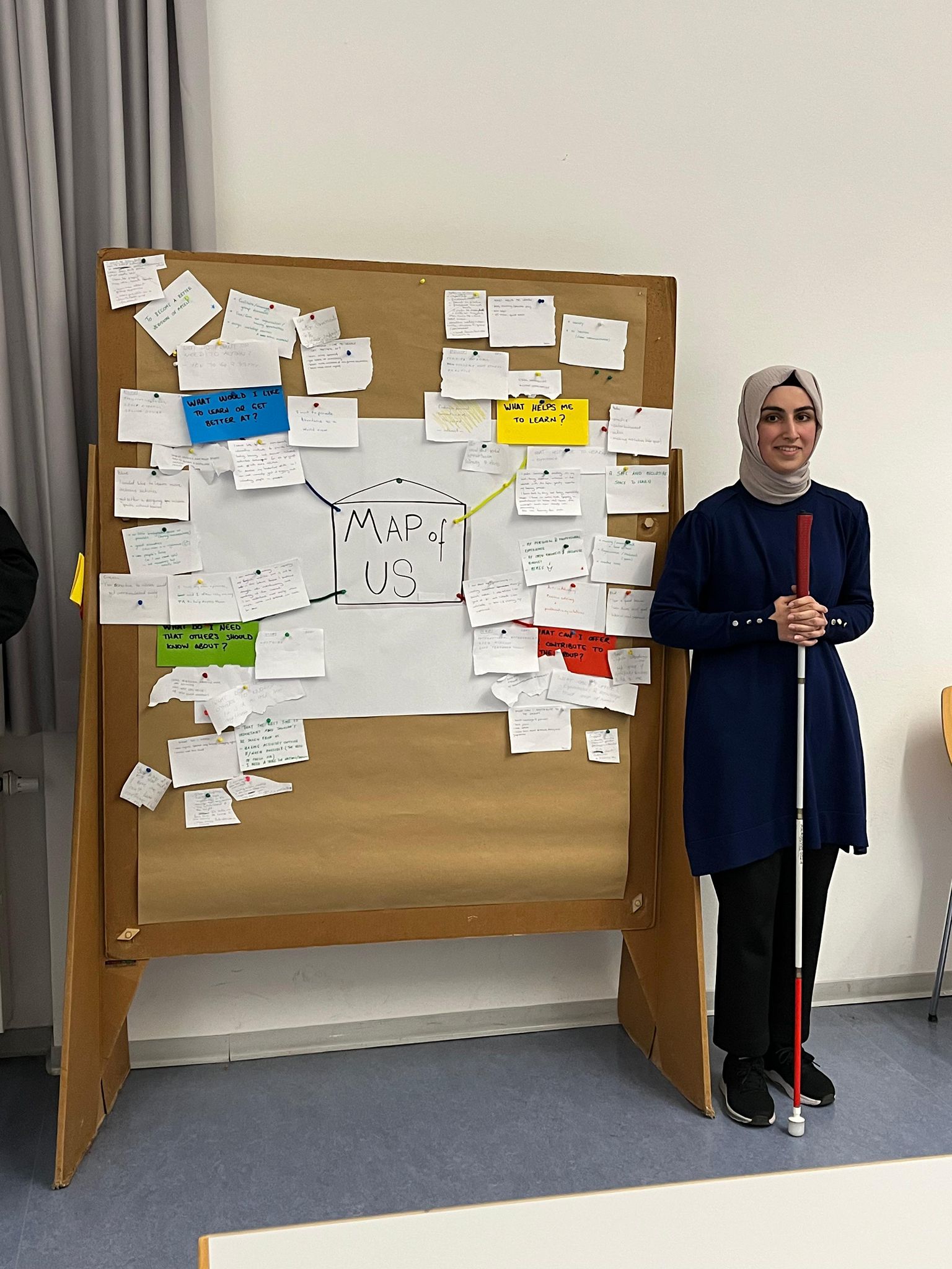 Fotoğrafta bir kadın, sınıf ya da eğitim ortamını andıran bir iç mekânda ayakta duruyor. Başörtüsü takıyor, lacivert uzun bir tunik ve siyah pantolon giymiş. Elinde kırmızı-beyaz renkte bir baston tutuyor. Yanında büyük bir pano var; üzerinde “MAP OF US” başlıklı bir kâğıt ve etrafına iğnelenmiş çok sayıda küçük not kâğıdı bulunuyor. Notlar renkli kâğıtlara yazılmış ve “What helps me to learn?”, “What would I like to learn or get better at?” gibi İngilizce sorular içeriyor. Arka planda gri perdeler ve açık renk duvarlar dikkat çekiyor. Fotoğraf genel olarak bir eğitim veya atölye çalışmasından bir anı yansıtıyor.