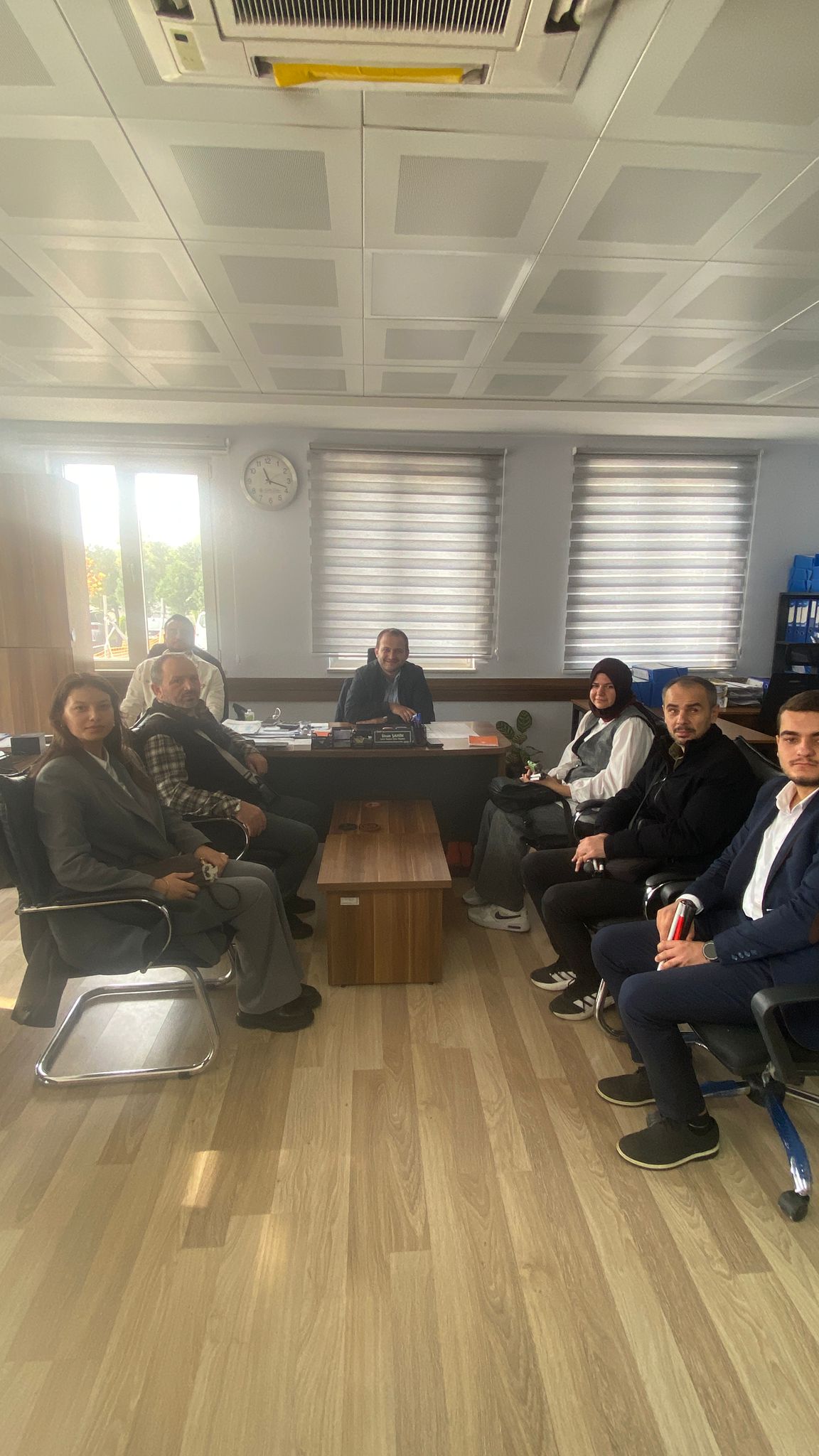 Fotoğrafta bir ofis ortamı görülüyor. Ortada bir masa ve masanın etrafında altı kişi bulunuyor. Masanın arkasında oturan kişi, müdür konumunda olduğu anlaşılan bir erkek. Masanın üzerinde çeşitli belgeler, dosyalar ve ofis malzemeleri yer alıyor. Masaya yakın oturan diğer beş kişi, ziyaretçi veya toplantı katılımcısı gibi görünüyor. Fotoğraftaki kişiler samimi bir ortamda, toplantı havasında poz vermiş durumda. Odada iki büyük pencere, duvarda bir saat ve tavanda kare panellerle kaplı bir tavan yapısı dikkat çekiyor. Aydınlık bir ortam mevcut, dışarıdan gelen gün ışığı odayı aydınlatıyor. Genel olarak, resmi ama sıcak bir görüşme anı yansıtılıyor.