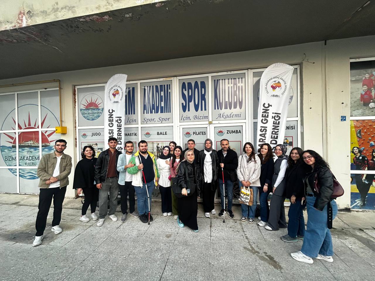 Fotoğrafta yaklaşık 15 kişilik bir grup binanın önünde toplu halde poz veriyor. Katılımcılar gülümseyerek kameraya bakıyorlar. Çoğunlukla genç yetişkinlerden oluşan grup, rahat ve günlük kıyafetler giymiş. Arka planda, binanın cephesinde “Bizim Akademi Spor Kulübü” yazılı tabelalar ve spor dallarını belirten (örneğin Judo, Jimnastik, Bale, Pilates, Zumba) yazılar görülüyor. Grupla birlikte iki adet beyaz renkli “Uluslararası Genç Aktivistler Derneği” yazılı dikey bayrak da yer almakta. Bu, fotoğrafın bir gönüllülük veya sosyal sorumluluk etkinliği kapsamında çekildiğini düşündürüyor. Zemin beton, duvarda ise kulübün logosu (güneş ve deniz figürleriyle) dikkat çekiyor. Katılımcıların arasında bir kişi elinde beyaz baston tutuyor, bu da kapsayıcı bir etkinlik olduğunu gösterebilir. Genel atmosfer samimi ve dayanışma ruhunu yansıtıyor.