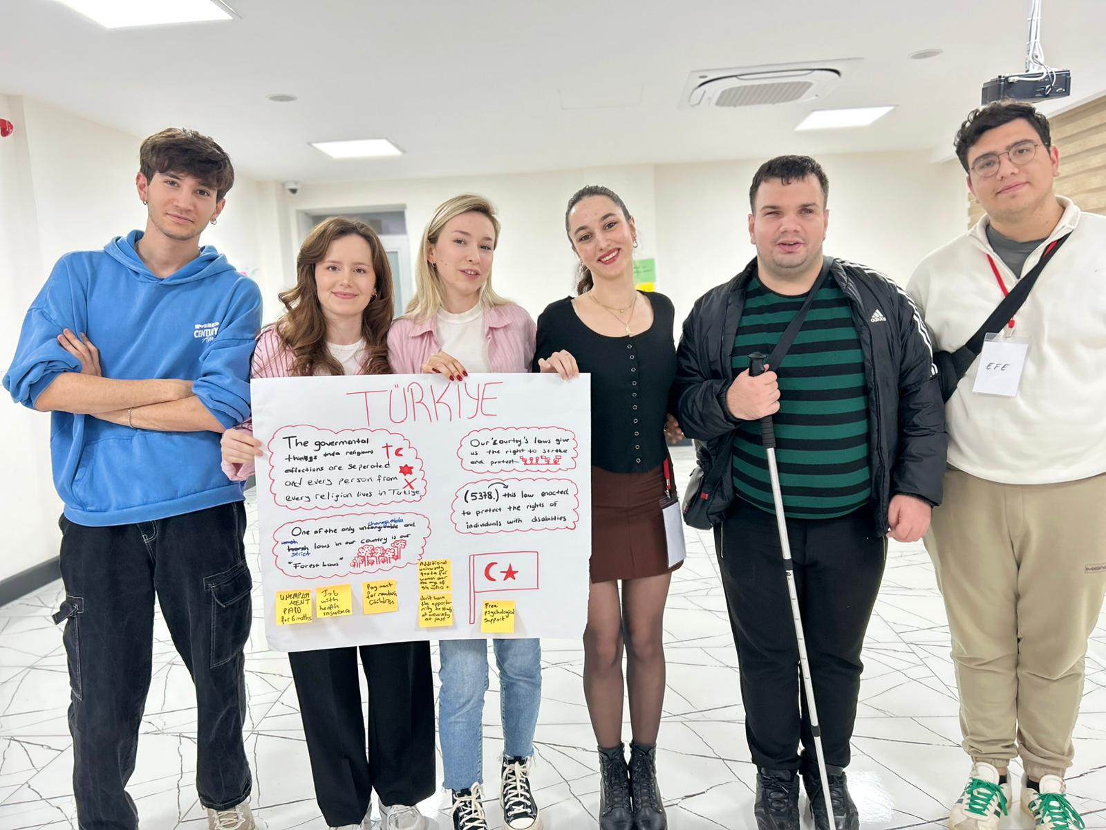 Fotoğrafta altı kişiden oluşan bir grup yan yana durarak kameraya poz veriyor. Hepsi kapalı bir toplantı veya eğitim salonunda, beyaz ışığın hâkim olduğu bir ortamda bulunuyor. Zeminde beyaz mermer desenli bir zemin var. Soldan sağa doğru: İlk kişi, mavi bir kapüşonlu sweatshirti ve siyah pantolonu ile rahat bir tarzda duruyor; kollarını hafifçe kavuşturmuş. İkinci kişi, dalgalı kahverengi saçlı, açık renk bir tişört ve beyaz tonlarında bir gömlek giymiş; gülümseyerek poz veriyor. Üçüncü kişi, sarı saçlı, pembe çizgili bir gömlek ve beyaz bir tişörtle fotoğrafta; elini ortadaki afişe uzatmış. Dördüncü kişi, siyah uzun kollu bir üst ve kahverengi etek giymiş; elinde “Türkiye” yazılı büyük bir poster tutuyor. Poster üzerinde İngilizce kısa açıklamalar, çizimler ve kırmızı renkle vurgulanmış notlar yer alıyor. Beşinci kişi, siyah-yeşil çizgili bir kazak ve siyah mont giymiş; elinde beyaz bir baston tutuyor ve gülümsüyor. Altıncı kişi, açık renk bir sweatshirt ve bej pantolon giymiş; boynunda isim kartı bulunuyor. Poster, Türkiye’deki mevzuatlar, haklar ve erişilebilirlik konularıyla ilgili İngilizce bilgiler içeriyor. Grup, bir proje sunumu ya da çalıştay faaliyeti kapsamında birlikte hazırladıkları çıktıyı sergiliyormuş gibi görünüyor. Genel atmosfer sıcak, katılımcı ve işbirliğine dayalı.