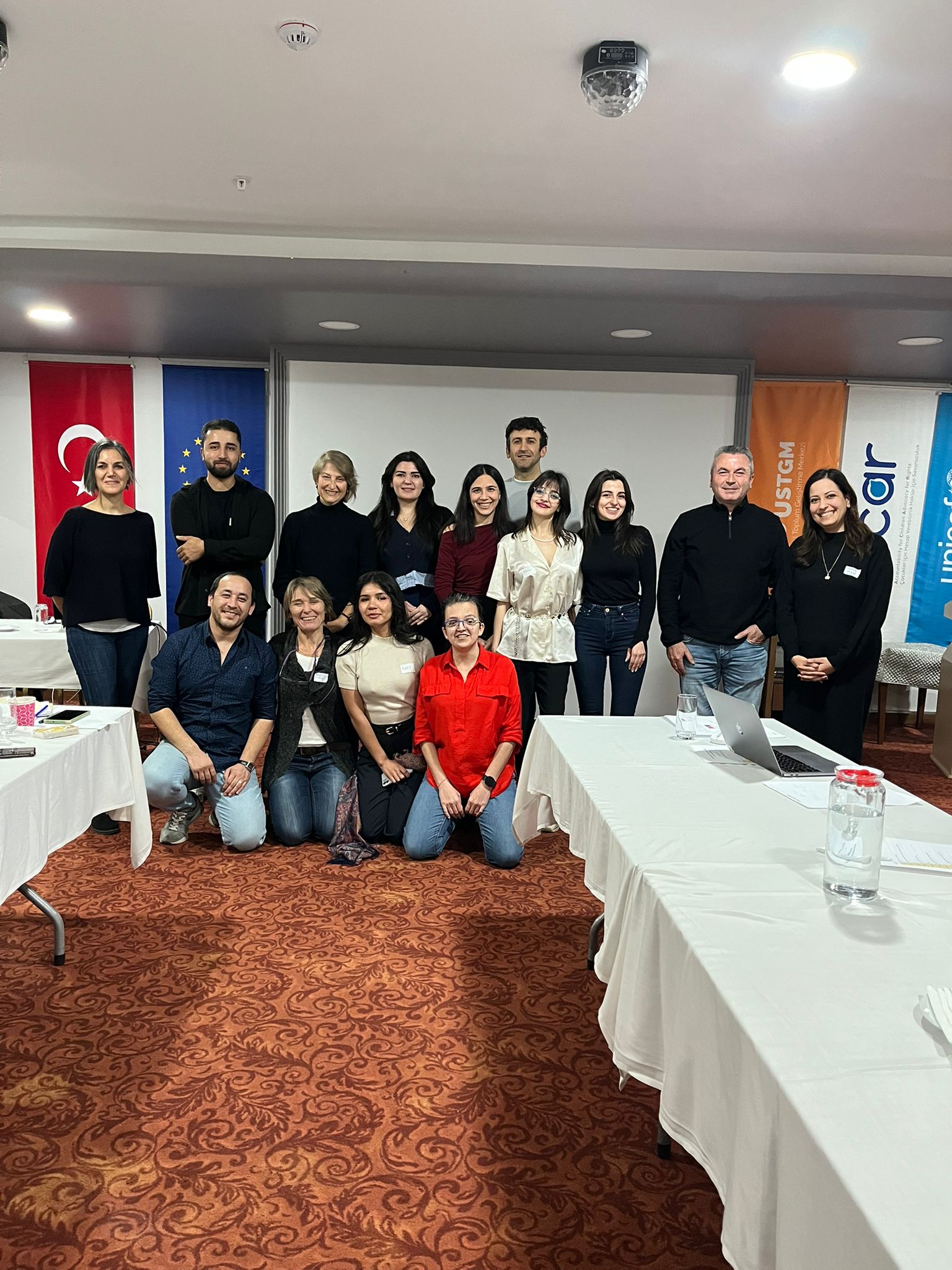 Fotoğrafta, bir toplantı veya eğitim etkinliği sonrasında çekilmiş gibi görünen kalabalık bir grup yer alıyor. Toplamda 15 kişi, iki sırada poz veriyor: arka sırada ayakta duranlar, ön sırada ise diz çökmüş veya yere oturmuş olanlar bulunuyor. Grup, gülümseyerek kameraya bakıyor ve genel atmosfer sıcak, samimi ve profesyonel bir birliktelik hissi veriyor. Arka planda, bir projeyle veya kurumla bağlantılı olduğu anlaşılan Türkiye bayrağı, Avrupa Birliği bayrağı ve yan tarafta “UNICEF” yazılı bir afiş görünür durumda. Bu da fotoğrafın uluslararası veya çok paydaşlı bir etkinlik kapsamında çekildiğini düşündürüyor. Katılımcıların çoğu sade ve şık kıyafetler giymiş; siyah tonlar ağırlıkta. Ortam bir otel konferans salonunu andırıyor: kırmızı desenli halı, beyaz örtülü toplantı masaları, sunum perdesi ve tavan aydınlatmalarıyla birlikte oldukça düzenli bir çalışma alanı görülüyor. Ön taraftaki masalarda su şişesi, belgeler ve bir dizüstü bilgisayar yer alıyor; bu da fotoğrafın bir oturum veya atölye sonrasında çekildiğini gösteriyor. Genel olarak fotoğraf; işbirliği, ekip ruhu ve profesyonel bir çalışma ortamının sıcak bir hatırası gibi görünüyor.