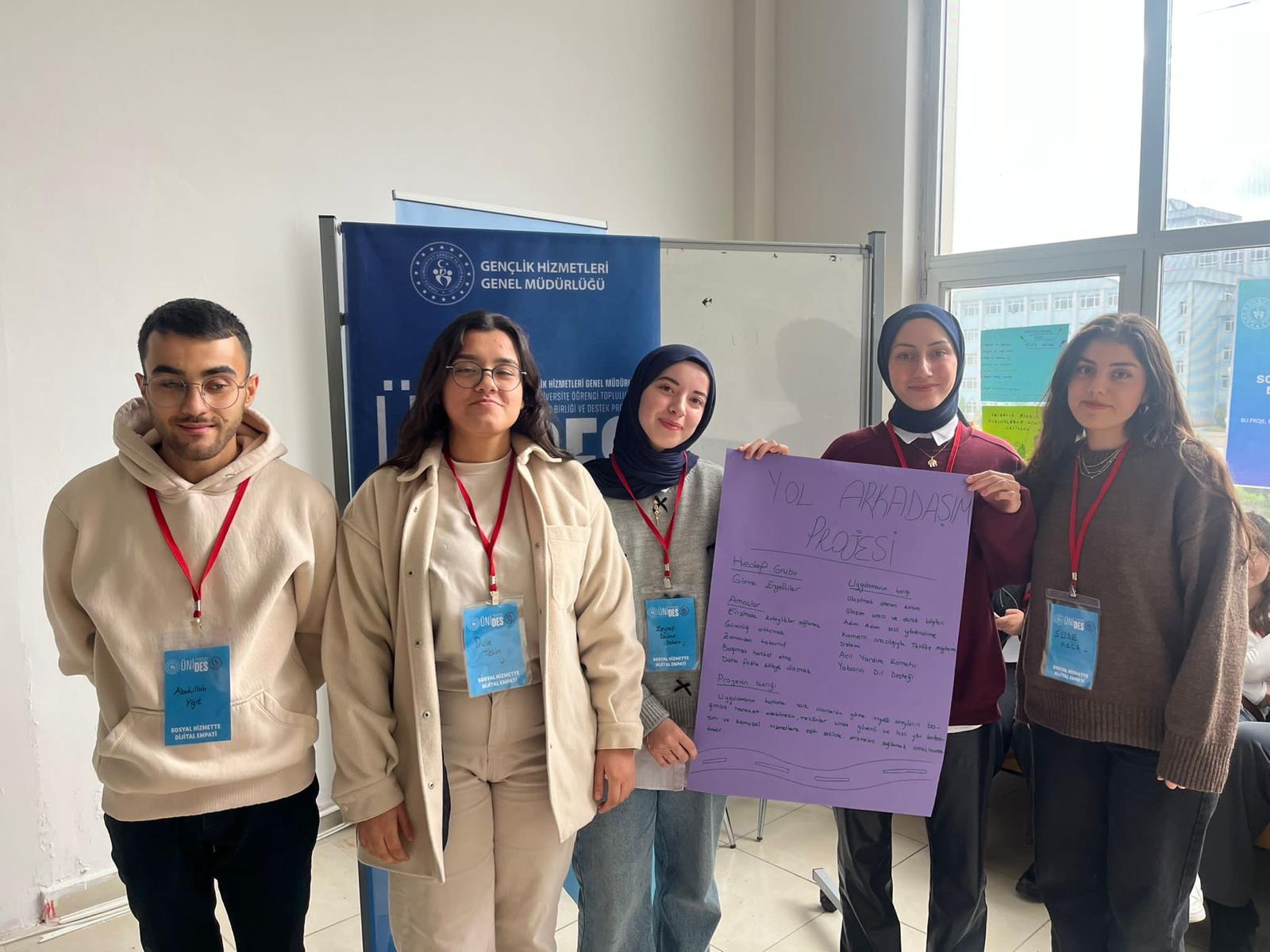 Fotoğrafta bir grup genç ayakta yan yana duruyor. Beş kişi var ve hepsi boyunlarında kırmızı ipli isimlik kartları taşıyor. Arka planda Gençlik Hizmetleri Genel Müdürlüğü yazılı bir banner ve beyaz bir tahta görülüyor. Mekân kapalı bir salon gibi görünüyor; sağ tarafta büyük bir pencere var ve dışarıda binalar seçiliyor. Soldan sağa doğru: İlk genç, bej hoodie giyiyor, gözlük takıyor ve gülümsüyor. İkinci genç yine bej tonlarında bir kombin giymiş, gözlük takıyor ve rahat bir şekilde poz veriyor. Ortadaki genç lacivert bir şal takmış, gri bir kazak giyiyor ve gülümsüyor. Dördüncü genç bordo bir kazak giymiş, lacivert bir şal takıyor ve elinde mor renkli büyük bir poster tutuyor. Sağdaki genç kahverengi bir kazak giymiş ve hafifçe gülümsüyor. Dördüncü ve beşinci kişi, üzerinde "Yol Arkadaşım Projesi" yazan mor renkli bir karton/poster tutuyor. Poster üzerinde proje hedef grubu, amaçlar ve uygulama adımlarına dair el yazısıyla maddeler yer alıyor. Genel atmosfer pozitif, ekip çalışması ve proje sunumu havası hâkim.